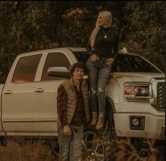 two people sitting on the hood of a white pickup truck in front of a wooded area