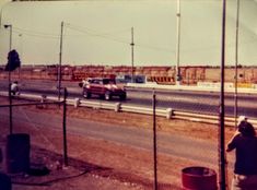 two cars driving down the road next to a fence and some people standing on the side of the road