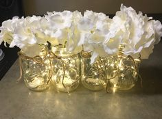 some white flowers are in glass vases with string lights on the bottom one is filled with water