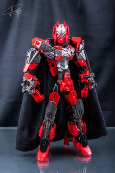 a red and black robot with a cape on it's head, standing in front of a black background