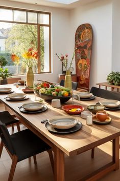 a dining room table set with plates, bowls and fruit in front of a large window