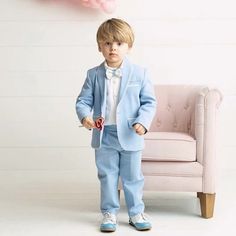 a little boy in a blue suit and bow tie standing next to a pink chair