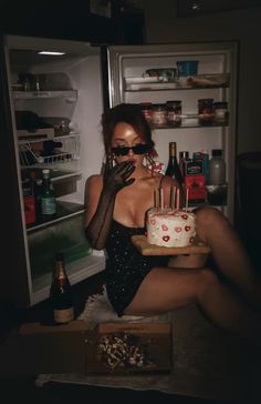 a woman sitting in front of an open refrigerator holding a cake with candles on it