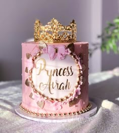 a pink princess cake with gold decorations and a tiara on top is sitting on a table