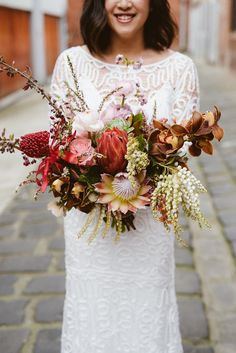 a woman in a white dress holding a bouquet of flowers and greenery on the street