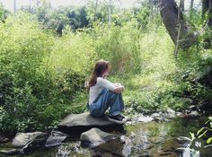 a woman sitting on top of a rock next to a stream in the forest with tall grass