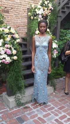 a woman in a blue dress standing next to some flowers and plants on a brick walkway