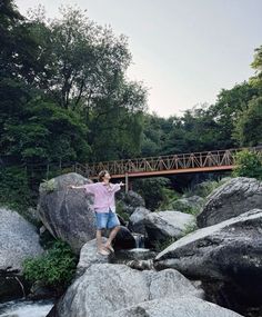 a man standing on top of a large rock next to a river with a bridge in the background