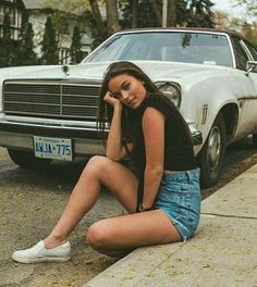 a young woman sitting on the curb next to a white car and holding her head