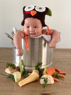 a baby wearing an owl hat sitting in a pot with carrots and celery