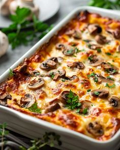 a casserole dish with mushrooms and cheese on top, surrounded by fresh herbs