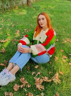 a woman sitting on the grass with her legs crossed and looking at the camera while wearing a multicolored sweater