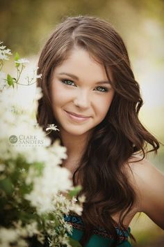 a girl with long brown hair and blue eyes smiles at the camera in front of white flowers