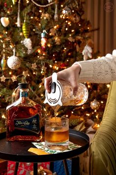 a person pouring an alcoholic drink into a glass on a table next to a christmas tree