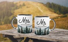 two coffee mugs sitting on top of a wooden table with mountains in the background