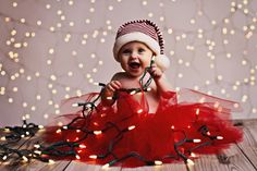 a baby wearing a red tutu while sitting on a wooden floor with christmas lights