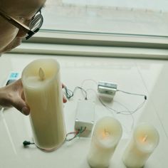 a man holding a lit candle in front of three smaller candles on a table next to a window