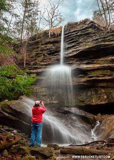 a person standing in front of a waterfall taking a photo with their cell phone while wearing a red jacket