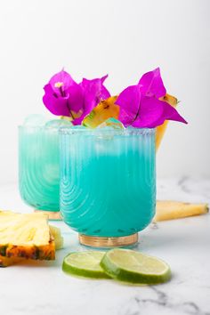 two glasses filled with blue liquid next to slices of pineapple and purple orchids