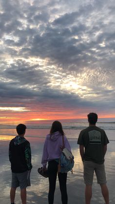 three people are standing on the beach watching the sun rise over the water and clouds