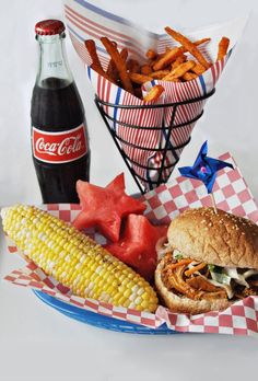 a plate with a sandwich, watermelon and corn on the cob next to a bottle of coca cola