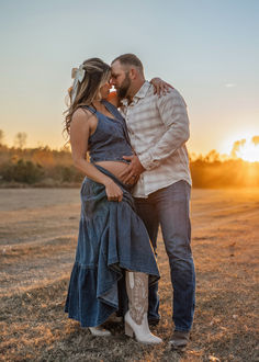 a man and woman standing next to each other in a field with the sun setting behind them