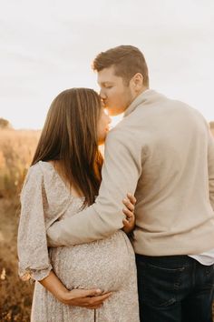 a pregnant couple cuddles and kisses in an open field at sunset with the sun shining on them