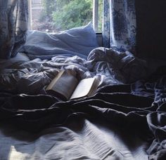 an open book laying on top of a bed in front of a window with curtains