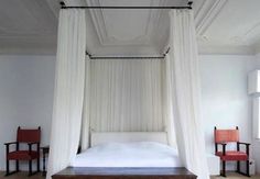 a canopy bed with white drapes over it in a room filled with wooden floors and red chairs