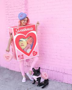 a woman in pink is holding up a sign with two hearts on it and a black dog sitting next to her