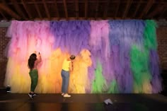 two people are standing in front of a large multicolored wall that is being painted