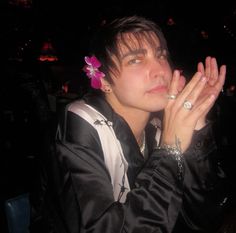 a young man sitting at a table with his hands together and wearing a flower in his hair