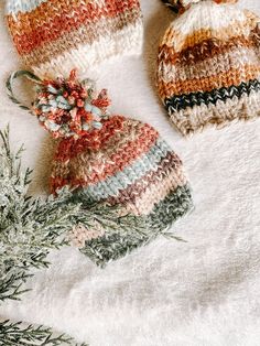 two knitted hats sitting on top of a white blanket next to a pine tree