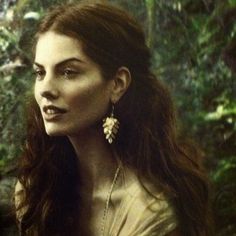 a woman with long brown hair standing in front of some trees and bushes wearing earrings