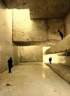 two people are walking through a tunnel with concrete walls and cement flooring, while one person is silhouetted in the distance