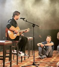 two men sitting on stools while one man plays the guitar and another sings into a microphone