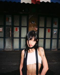 a woman with black hair and tattoos standing in front of a storefront wearing suspenders