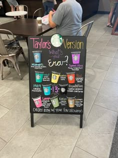 a menu board with drinks on it in a restaurant or bar area that has people sitting at tables and standing around