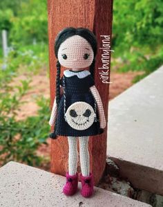 a crocheted doll wearing a black dress and pink boots stands next to a wooden post