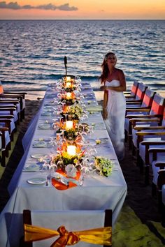 a woman standing next to a long table covered in flowers and candles at the beach