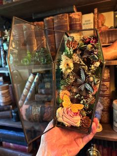 a person holding up a glass box with flowers and butterflies on it in front of bookshelves