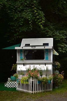 a small white house with flowers and plants on the front porch, next to a fence