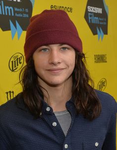 a woman with long hair wearing a beanie smiles at the camera while standing in front of a yellow background