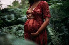 a pregnant woman in a red dress is standing among the green plants and trees with her hands on her stomach