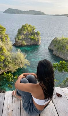 a woman sitting on top of a wooden dock next to the ocean and trees in the background