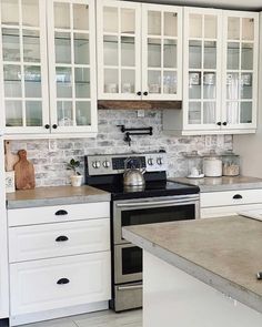 An opulent white farmhouse kitchen designed with white windowpane cabinetry with white grid patterns and white cabinets with black hardware. A neutral, gray countertop is paired with a whitewashed, exposed brick backsplash. A large white kitchen island with a neutral countertop occupies the center of the room and light wood flooring runs throughout. Contemporary decor adorns the countertops and soft natural light illuminates the space...   Image: thishouseofsix