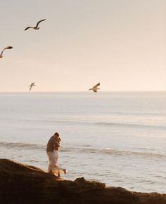 two people standing on the edge of a cliff by the ocean with seagulls flying overhead