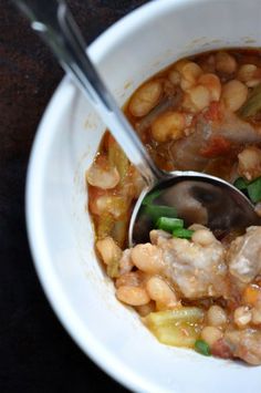 a white bowl filled with beans and meat next to a spoon on top of a table