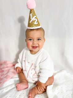 a baby wearing a birthday hat on top of a white blanket with pink pom poms