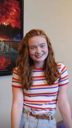 a girl with long red hair smiles in front of a movie poster on the wall